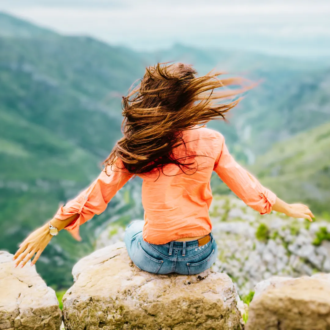 Back of woman sat on top of hill with arms wide, wind blowing behind her