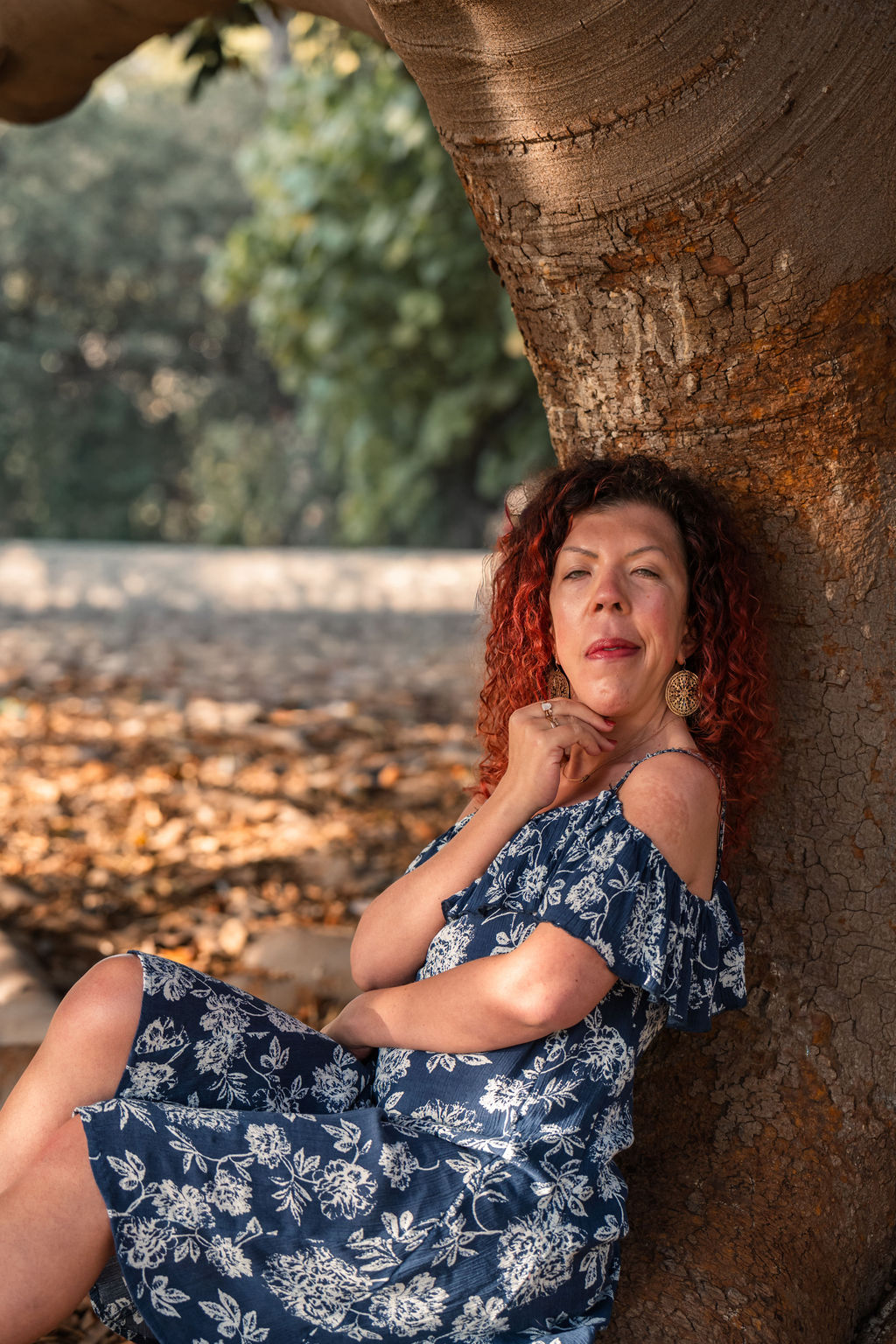 A grounded woman that can guide transformation safely is sitting on a root from a tree and leaning against the trunk. Gazing – transparent, opening, real.