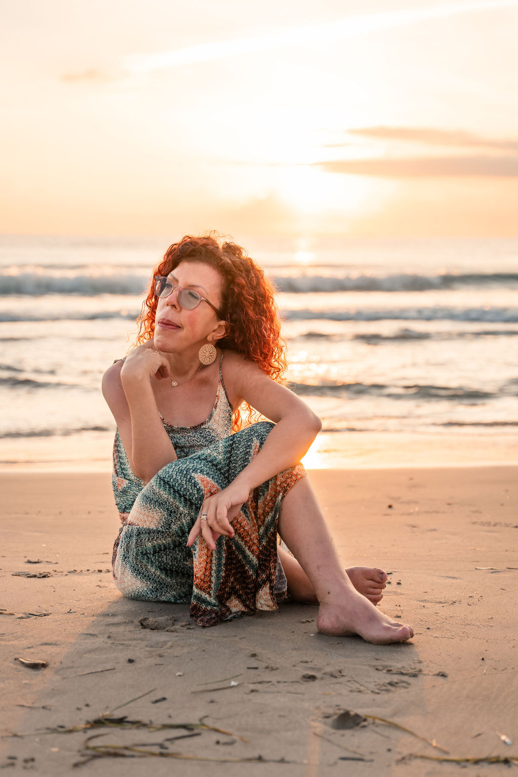 A contemplative woman sits on the beach. Rolling waves and a glowing sunrise in the background. A dress in pastel colors.