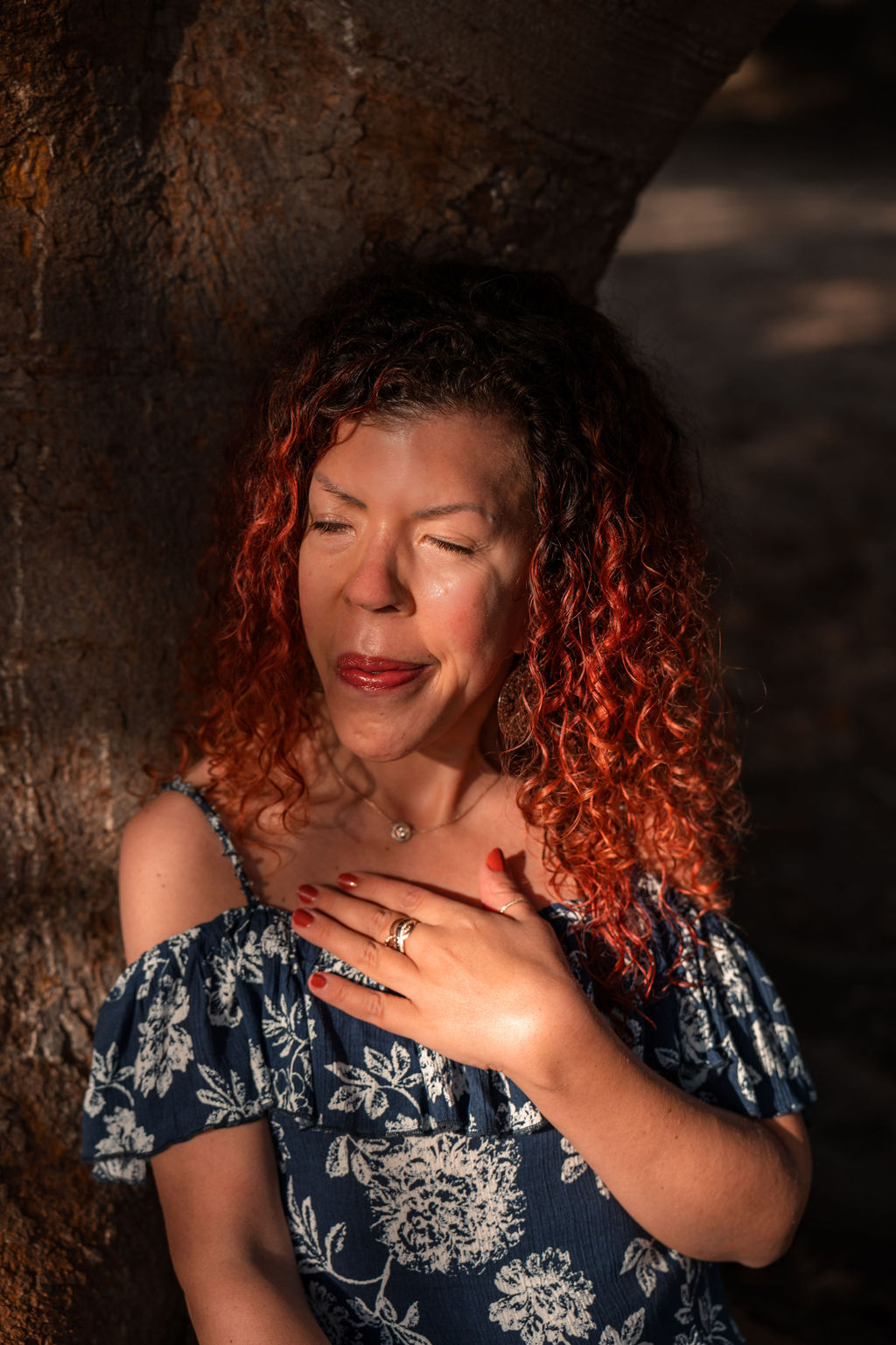 A grounded woman that can guide transformation safely is sitting on a root from a tree and leaning against the trunk. Eyes closed, sun shining on her face and hand on heart.