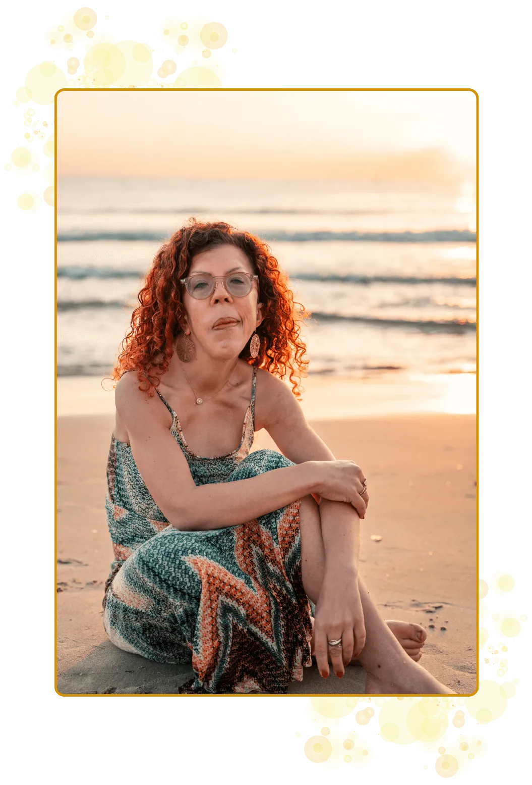 Smlende kvinne sitter på stranden med solens gylne lys og bølgeskvulp i bakgrunnen. Lang kjole i duse farge.