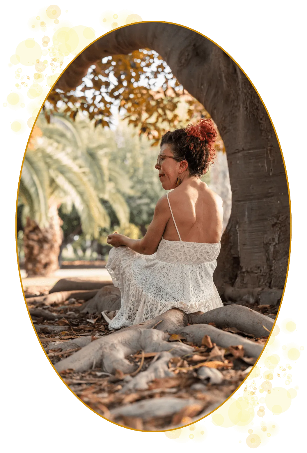 Kvinne med skoliose (skjev ryggrad), oppsatt hår, hvit kjole i bohemsk stil og sittende på et stort og kraftfullt tre sine røtter.