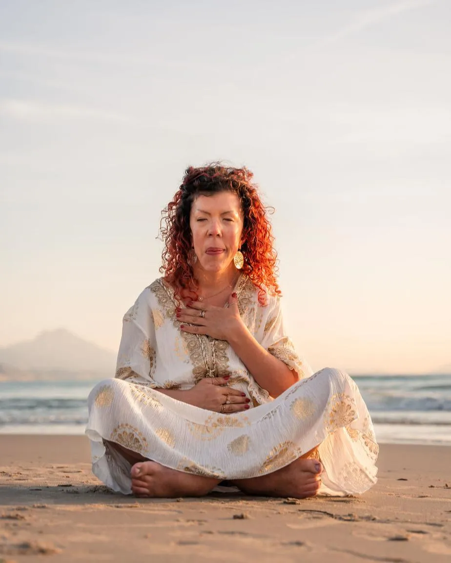 An embodied woman with a dress in white and gold. Sitting on the beach in sunrise. Red, curly hair. One hand on heart (heart chakra), one hand on stomach (sacral chackra).
