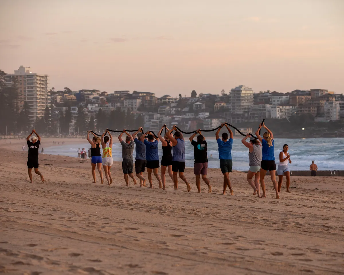 A man and a woman running away from the camera along a coastal pathway at sunrise or sunset, wearing black tank tops with MBHC logos. Beautiful ocean and headland in the background.