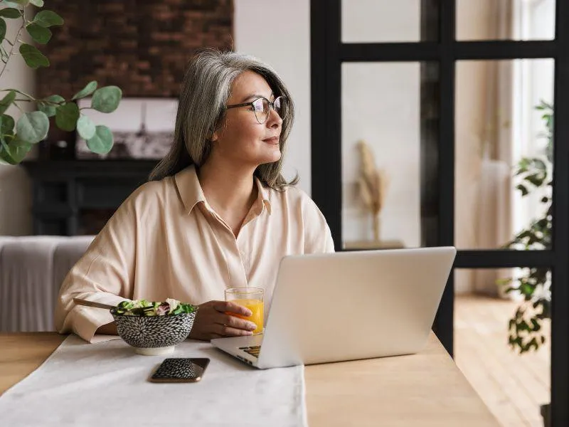 lady sat at desk with computer looking out o window with glass or orange juice in hand