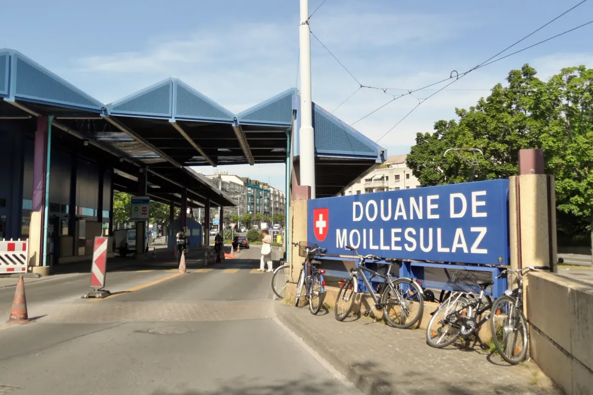 Panneau bleu « Douane de Moillesulaz » frappé de la croix suisse, devant l'auvent du poste frontière et des vélos stationnés