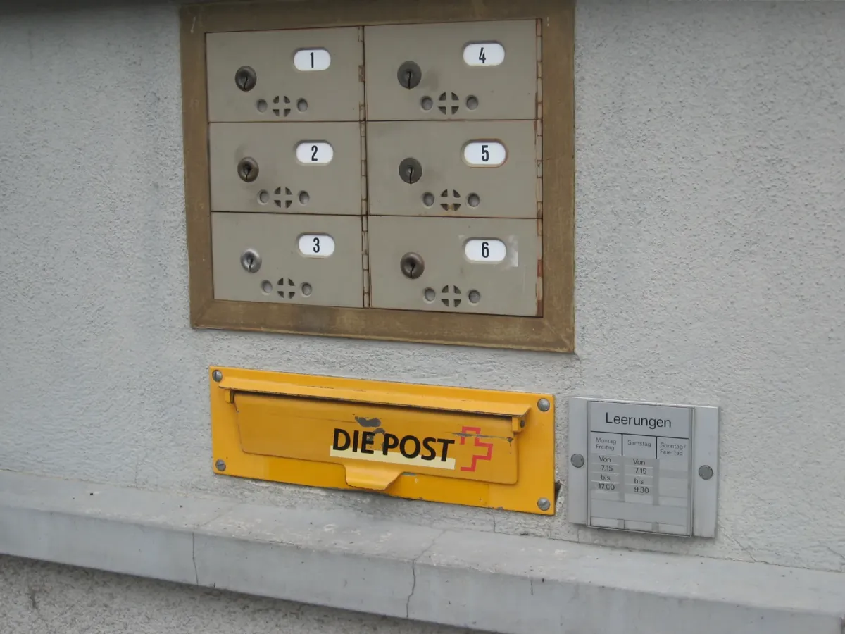 Boîte aux lettres jaune marquée Die Post, fixée au mur sous un bloc de six cases postales numérotées