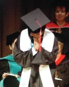 A graduate in a black gown and cap bows with hands together in gratitude.
