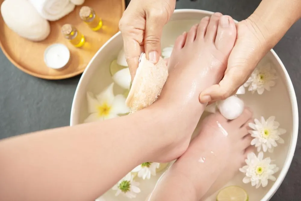 A serene and inviting pedicure scene where hands gently exfoliate a client's feet soaking in a bowl of warm water adorned with white flowers and lemon slices. The tranquil setting is enhanced by essential oil bottles, a soft towel, and a lit candle in the background, suggesting a relaxing and rejuvenating spa experience