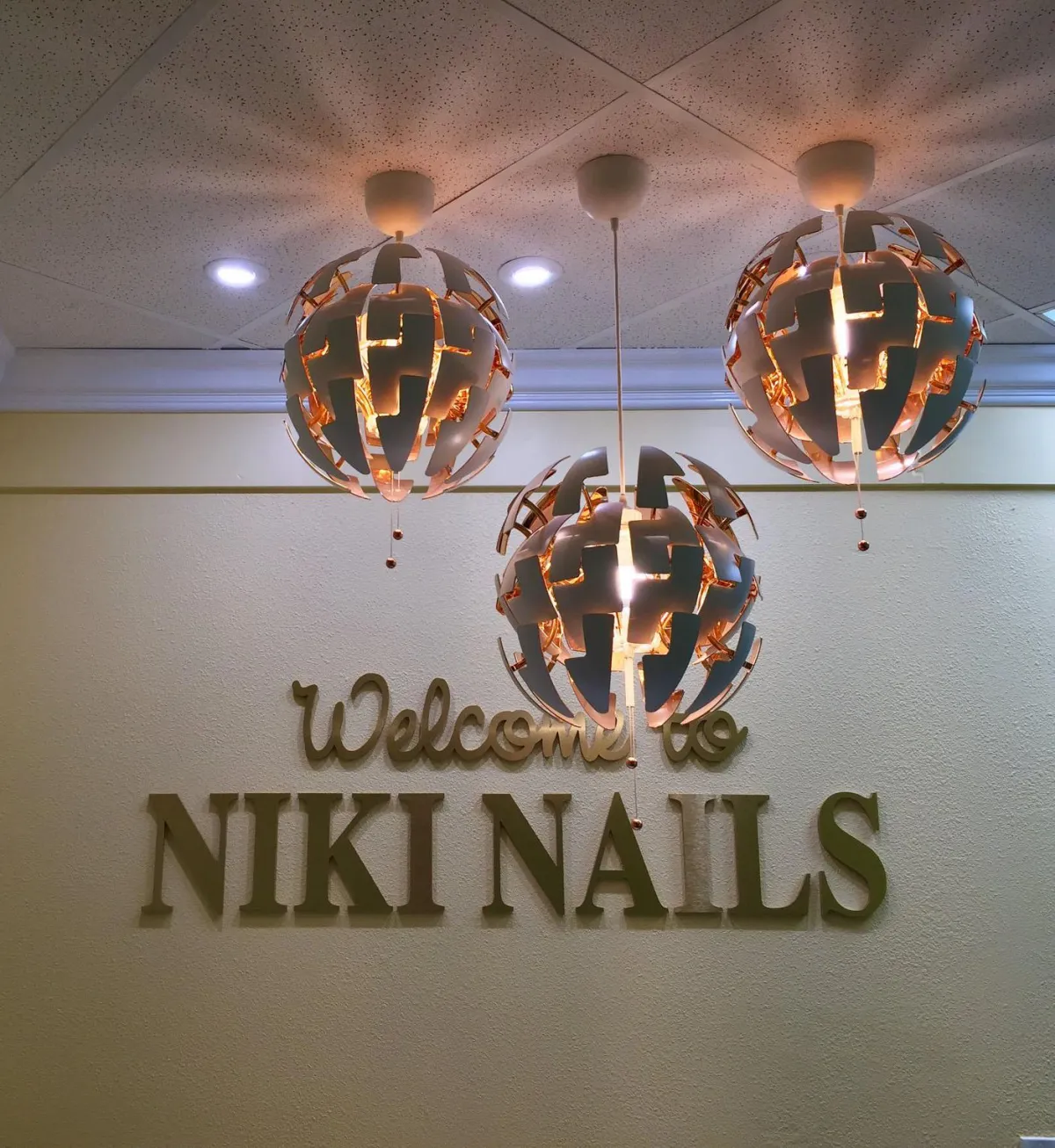 Interior view of a salon featuring three spherical pendant chandeliers with a geometric design and warm, glowing lights. Below the chandeliers, 'Welcome to NIKI NAILS' is written in elegant, capital, bronze letters on a textured off-white wall.