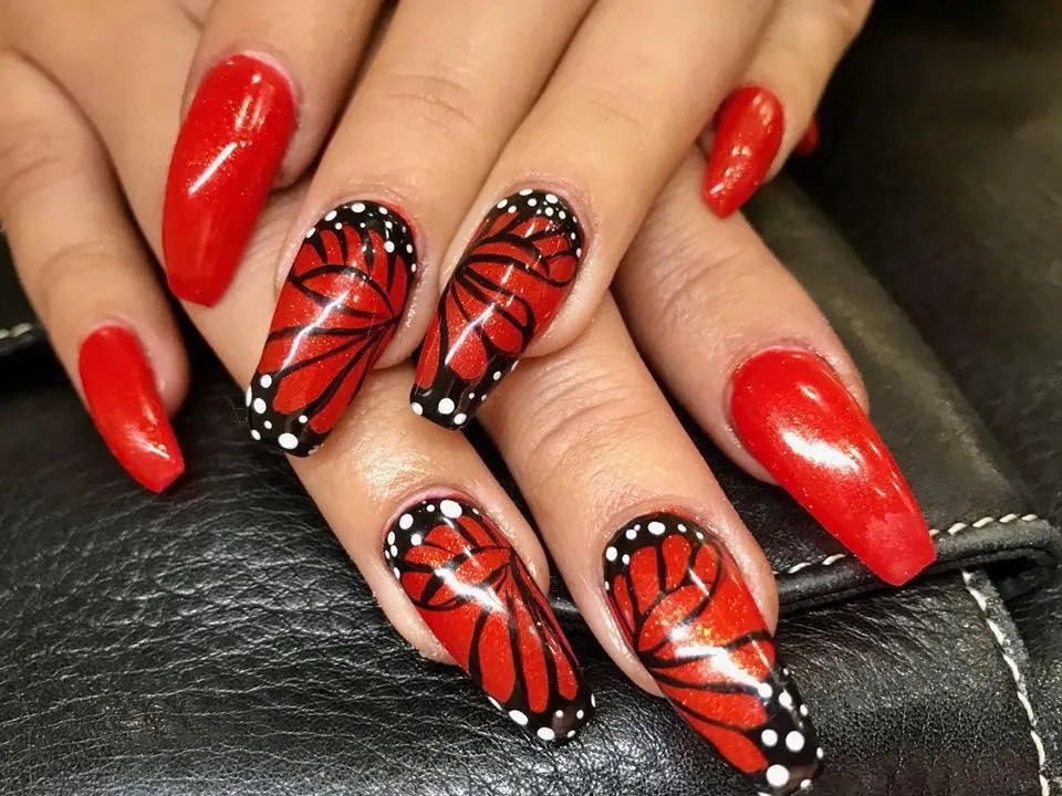 A close-up of a client's hand showcasing a striking nail art design. The nails are long and sculpted into a stiletto shape, painted in a glossy, vibrant red. Two of the nails on each hand feature a detailed depiction of butterfly wings with black and white accents, creating a bold, dramatic effect against the shiny red background.