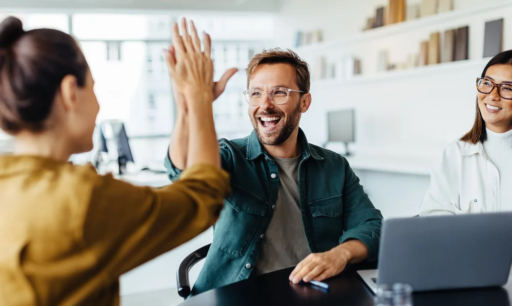 The image shows a group of three people in a modern office setting. The focus is on a man wearing glasses, smiling broadly as he shares a high-five with a colleague. The moment captures a sense of camaraderie and achievement