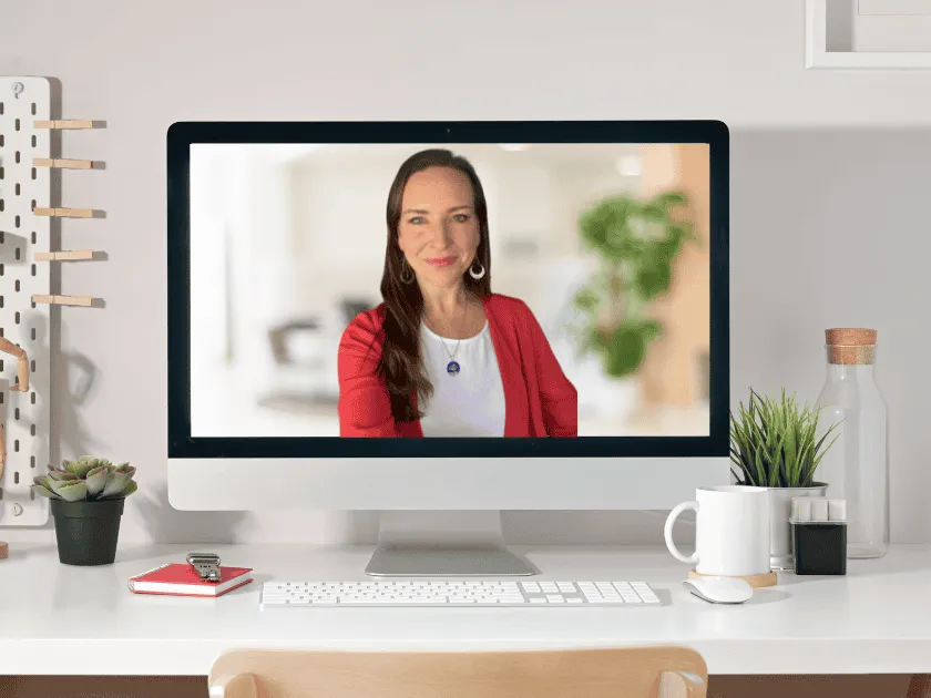 Computer monitor displaying a smiling woman in a red blazer and white top on a video call, set on a clean, modern desk with plants and office supplies.