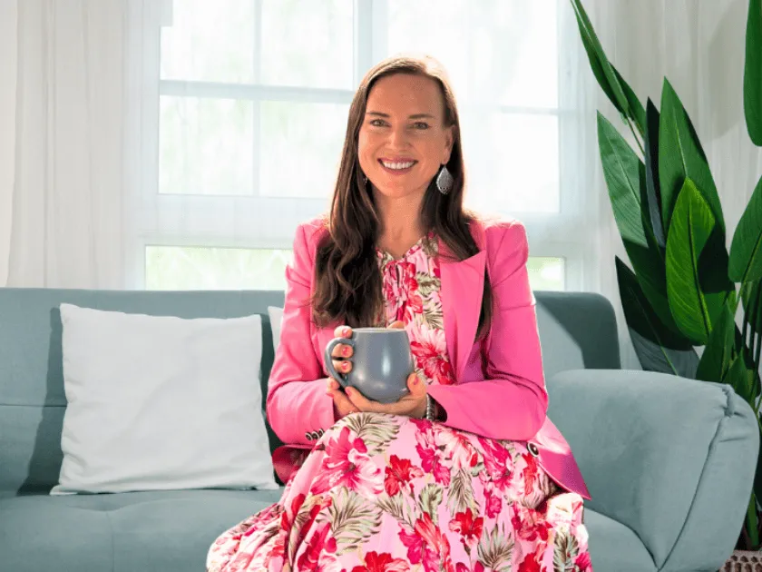 Smiling woman seated on a light blue sofa, wearing a pink blazer and floral dress, holding a gray mug in a bright, plant-filled room.