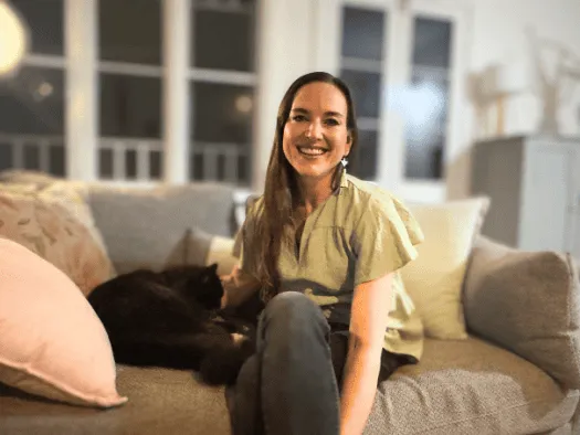 Woman smiling while sitting on a cozy sofa with a black cat resting beside her in a warmly lit living room.