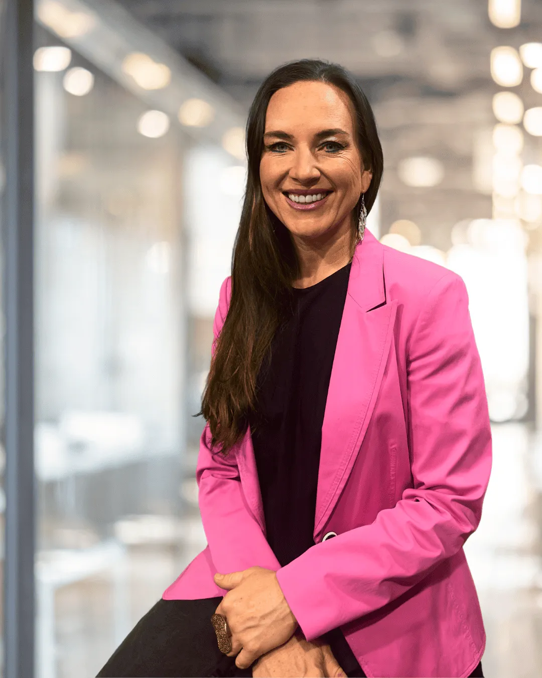 Smiling woman in a bright pink blazer, seated in a modern office with blurred lighting and a professional ambiance.