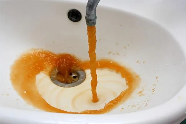 Orange-brown tap water flowing from a corroded galvanized pipe into a white bathroom sink.