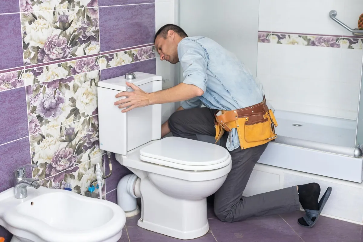 Technician examining a leaking toilet valve assembly in a tiled bathroom with visible water damage concerns.