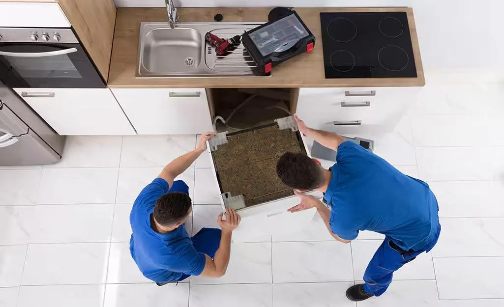 Plumbers installing a new dishwasher under kitchen counter, handling hookups and drainage.