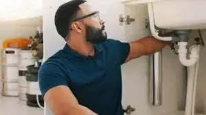 Plumber securing loose plastic P-trap under sink to fix odor and gas seep issues.
