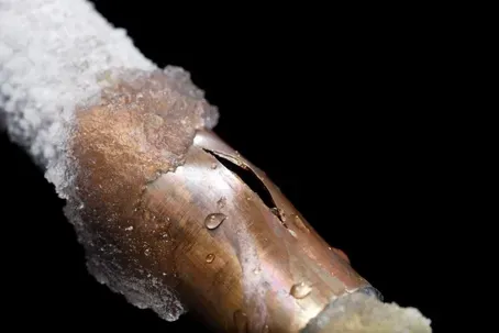 Copper water line cracked open from frost damage, covered in ice — a common winter pipe failure in basements and yards.