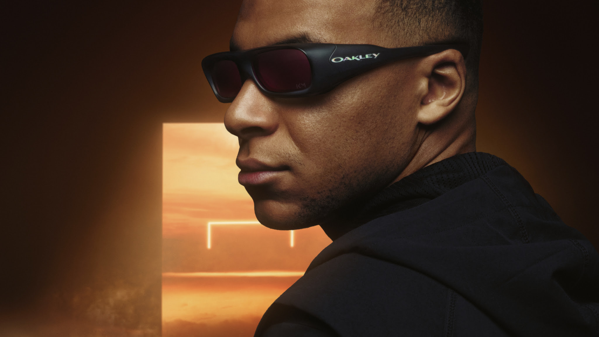 Man wearing black Oakley performance sunglasses against a dramatic orange background.