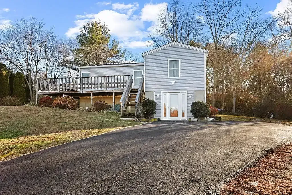 Completed home exterior remodel by KMAX Renovations in Guilford CT, showing new light gray vinyl siding, a custom wood deck with stairs, a modern glass front entry door, and a clean paved asphalt driveway.