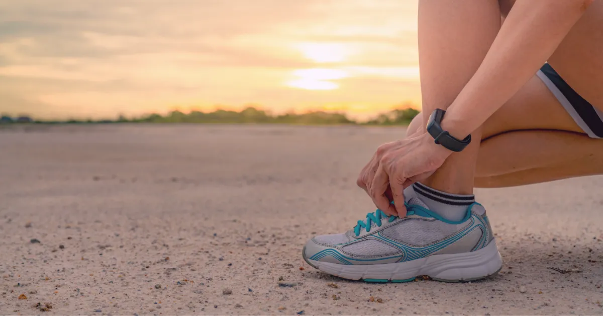 Woman tying her running shoe laces on a dirt path at sunrise, wearing a fitness tracker watch