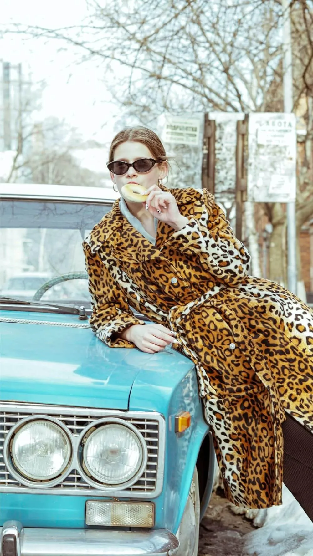 Woman in a leopard print coat leaning on an aqua old vehicle eating a bagel