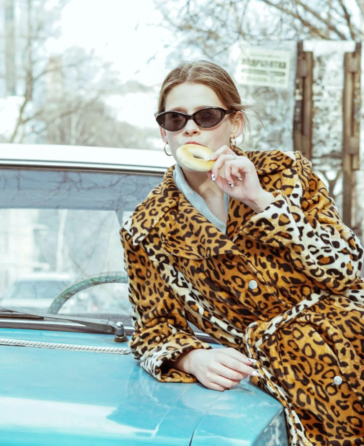 woman wearing sunglasses and leopard coat eating a bagel leaning on blue car