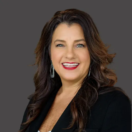A smiling Asian woman in her late 30s with long black hair, wearing a silk blouse and delicate gold necklace, standing beside a bookshelf filled with design books. The lighting is warm and inviting.