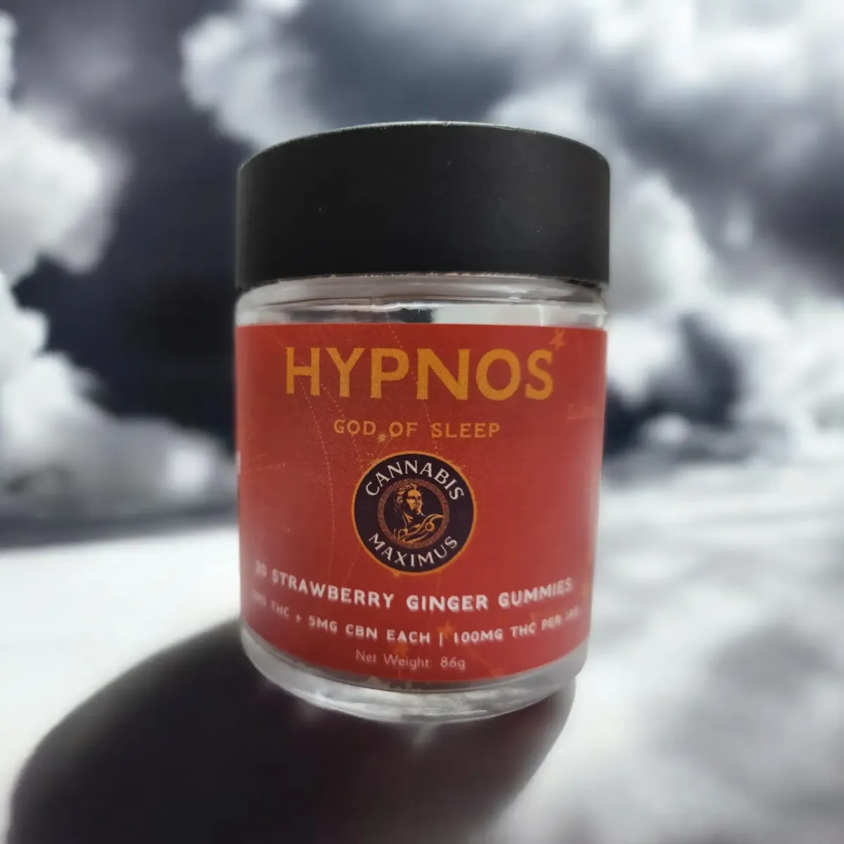 Cannabis Maximus Hypnos gummies jar with red label reading “God of Sleep” and “Strawberry Ginger Gummies” against a cloudy sky background
