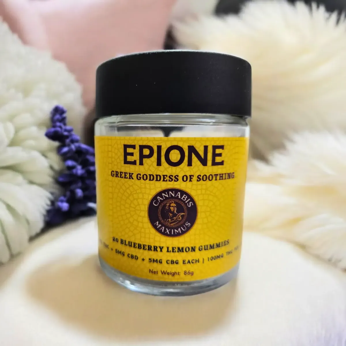 Cannabis Maximus Epione gummies jar with yellow label reading “Greek Goddess of Soothing” and “Blueberry Lemon Gummies” placed among soft pillows and flowers