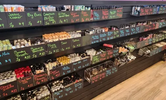 Cannabis products displayed on shelves inside Cannabis Maximus dispensary in Brattleboro VT showing flower jars, pre-rolls, concentrates, cartridges, and edibles with handwritten strain names and prices on black display boards
