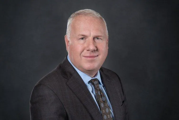 Professional headshot of Adam C. Rohrig, President and cofounder of Centurion Insurance Services, wearing a dark suit, light blue shirt, and patterned tie against a dark backdrop.