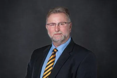 Professional headshot of Timothy J. Mitchell, GBDS, AIF®, Life & Retirement Plan Consultant at Centurion Insurance Services, wearing a dark suit, light blue shirt, and yellow-striped tie, posed against a dark background.