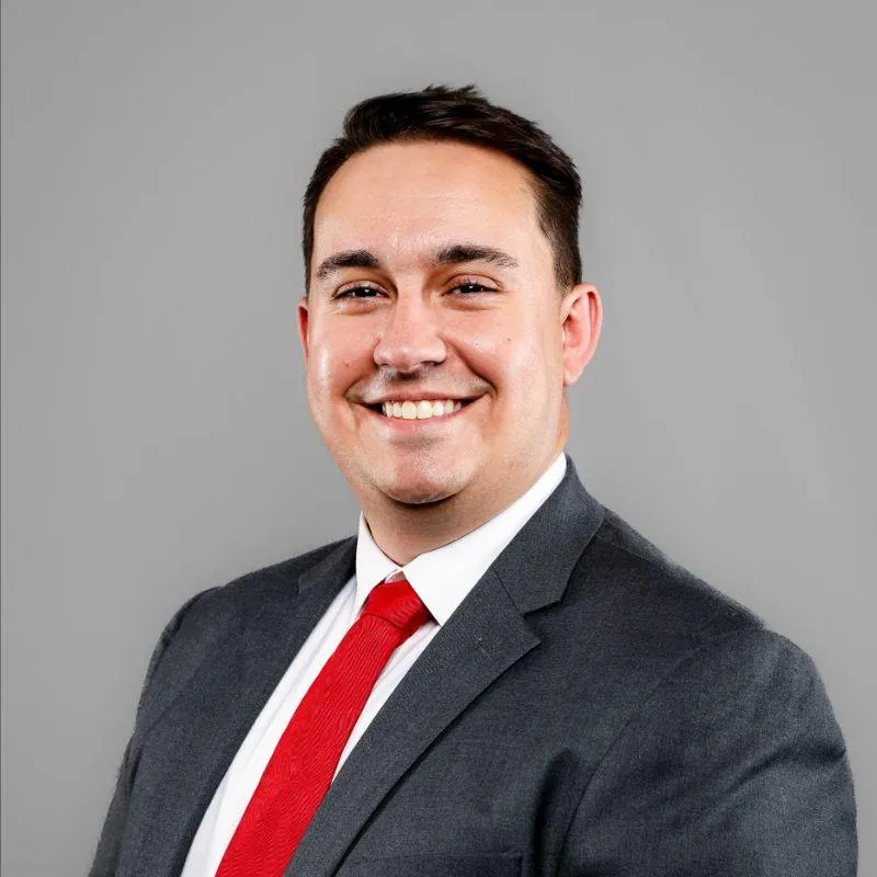 Professional headshot of Zack Cleary, REBC®, team member at Centurion Insurance Services, wearing a dark gray suit, white shirt, and red tie against a gray background.
