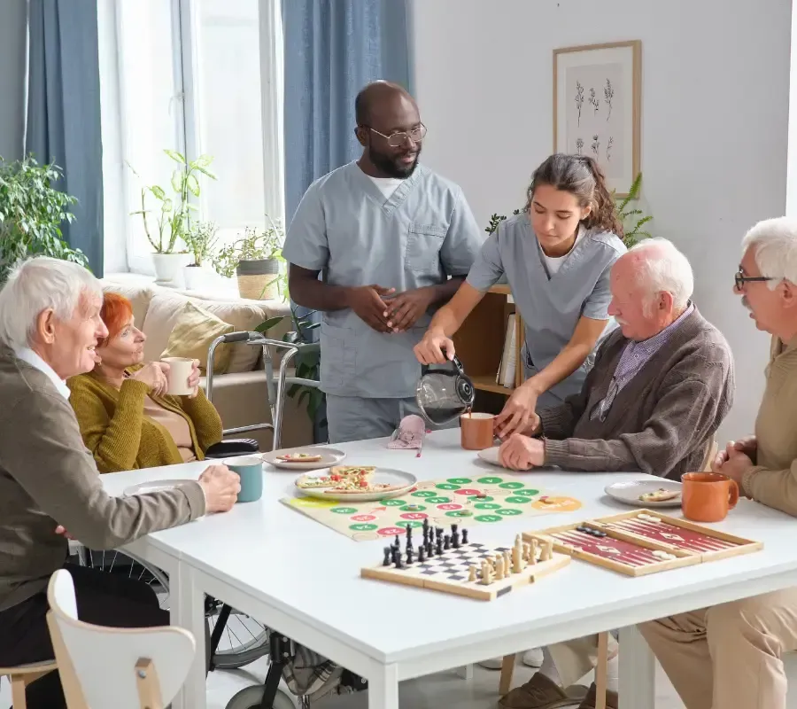 Caregivers serve drinks to older adults gathered around a table with board games in a bright assisted living common room.