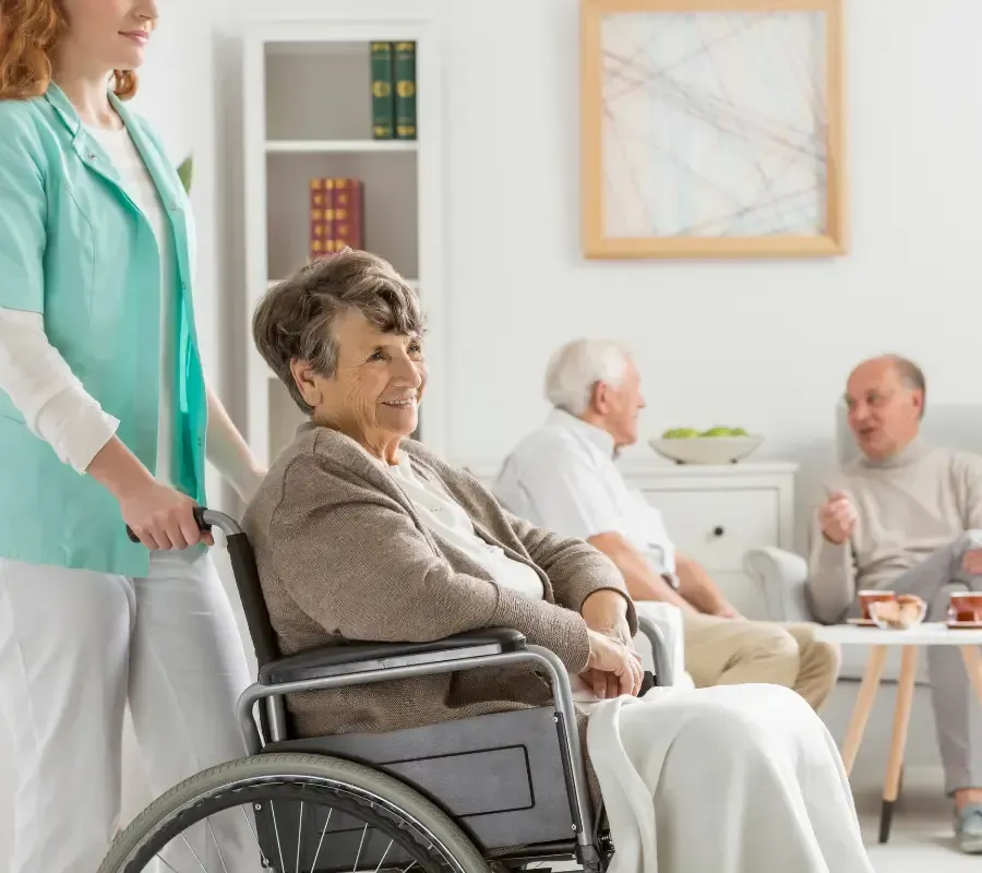 Caregiver standing beside an older woman seated in a wheelchair in a bright assisted living facility, with other residents talking in the background.