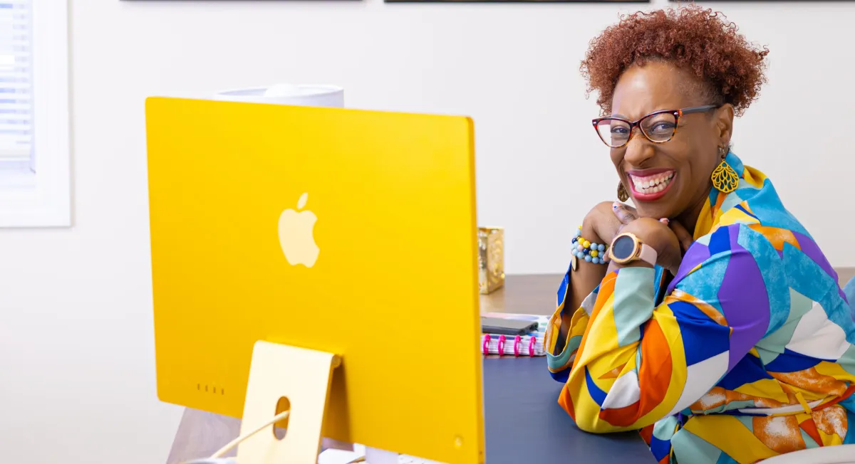 Dr. Natasha Booker smiling at her desk in front of a yellow computer — life coach and therapist for ambitious Black women focused on healing, leadership, and personal growth.