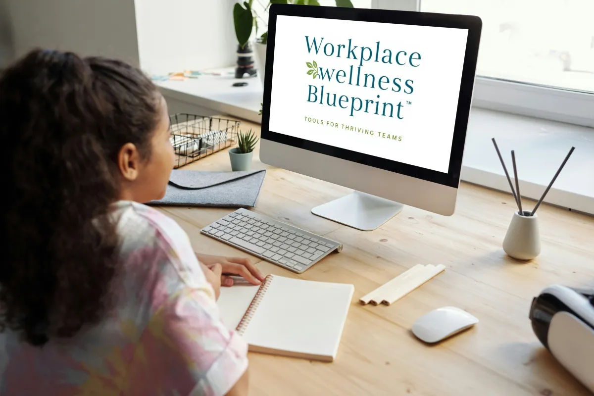 Woman sitting at a desk viewing “Workplace Wellness Blueprint” on a computer screen — a weekly newsletter for organizations focused on emotional wellness, leadership, and team development.