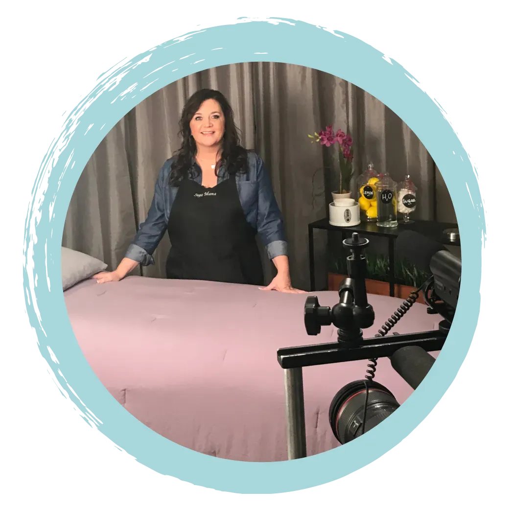 Woman in apron standing by a massage table, preparing for body sugaring training, with camera equipment and decorative plants in the background, emphasizing professional sugaring techniques.
