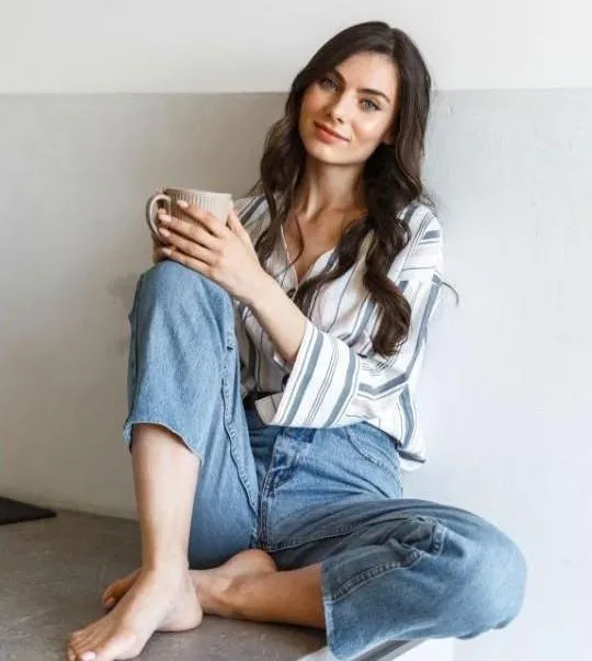 Woman sitting casually with a cup, wearing a striped shirt and relaxed jeans, embodying a welcoming atmosphere for beauty professionals in sugaring education.