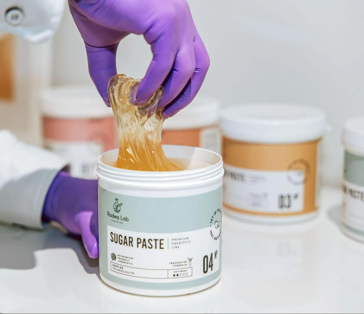 Hand reaching into a container of sugar paste, showcasing body sugaring technique for beauty treatments, with additional containers in the background.