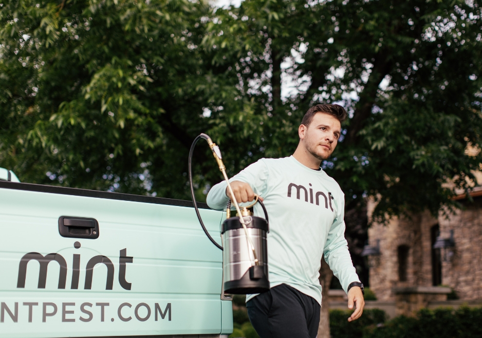 Mint Pest Control technician carrying treatment equipment beside a branded service vehicle