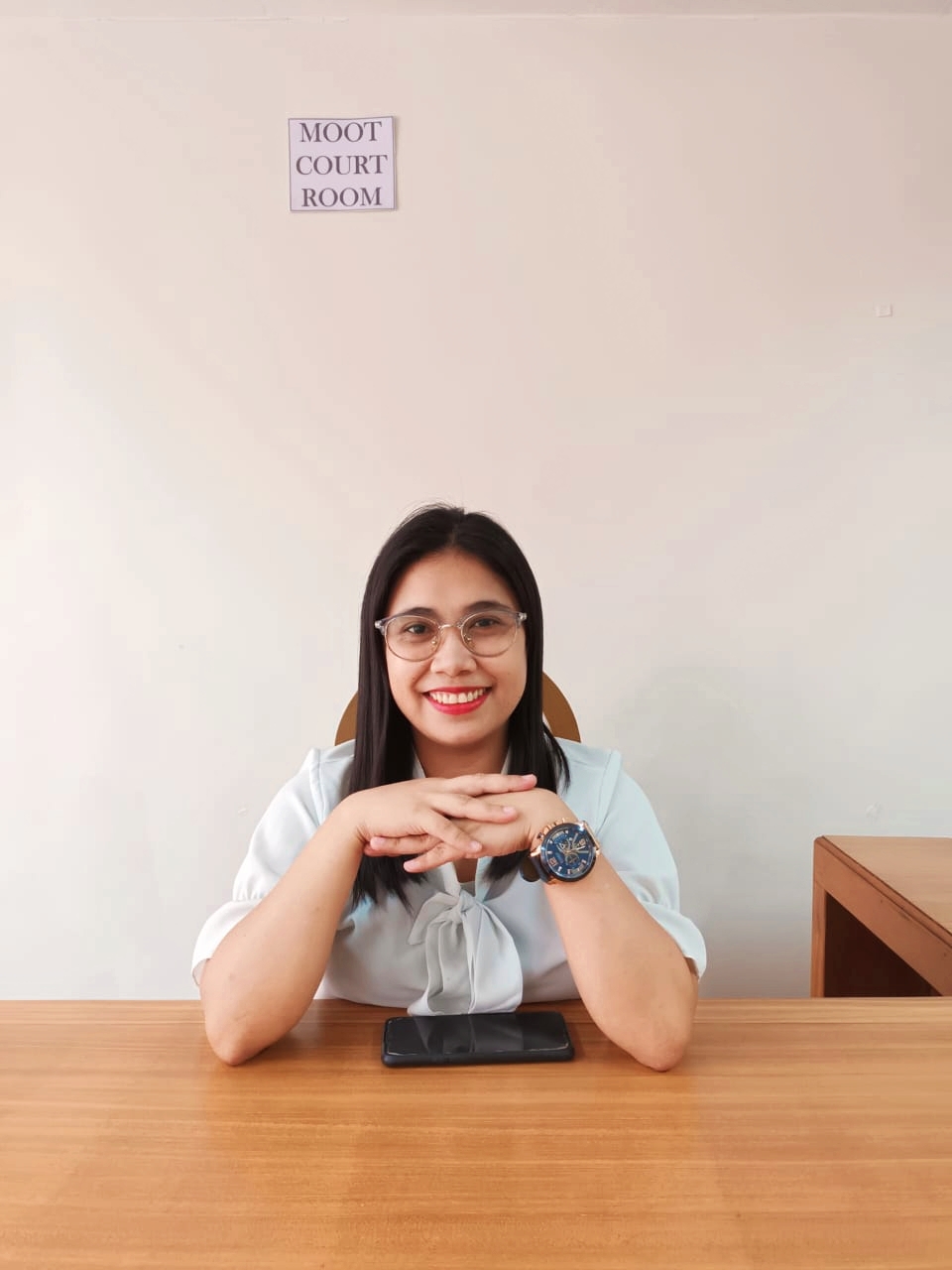 Smiling professional woman wearing glasses and a light blue blouse, seated at a wooden desk with hands folded, looking confident and approachable in a minimalist office setting