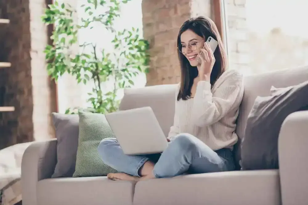 A woman is sitting on a couch, engaged in a phone conversation, and simultaneously operating a laptop for probate administration or trusts-related purposes.
