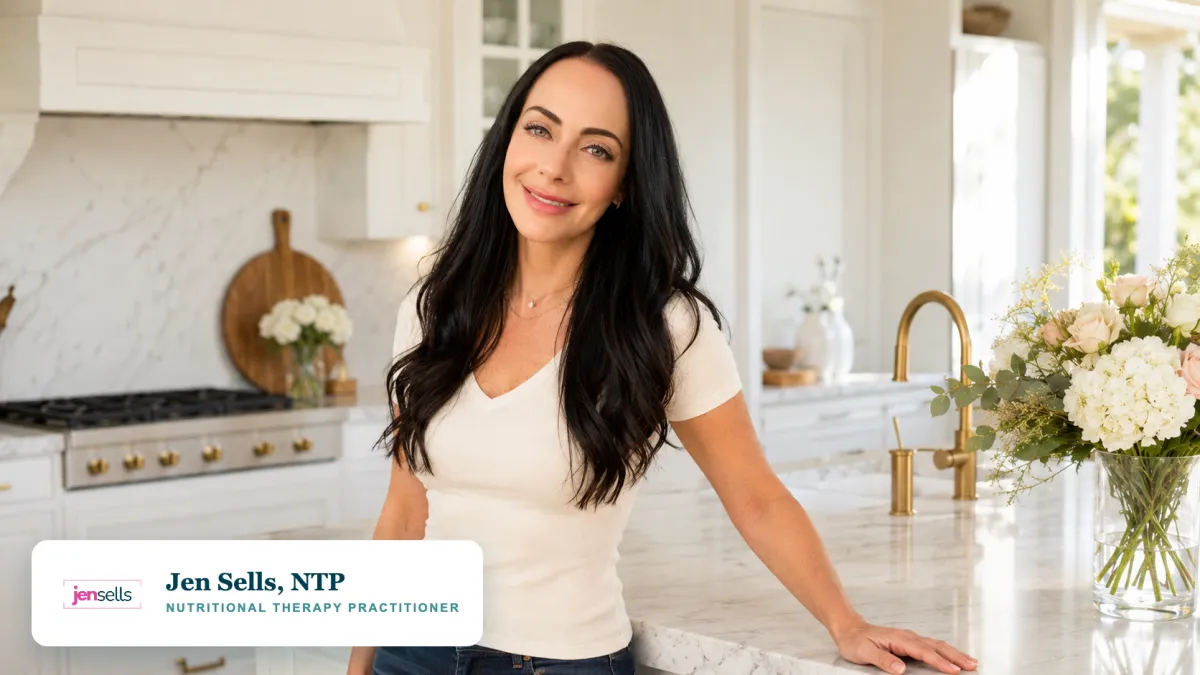 Jen Sells, Nutritional Therapy Practitioner, smiling in her bright white kitchen with marble counters and fresh flowers