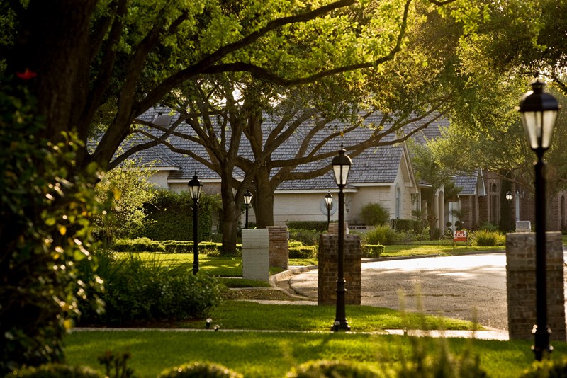 Tree-lined residential street with manicured lawns, black lamp posts, brick pillars, and homes with slate roofs in a well-established neighborhood