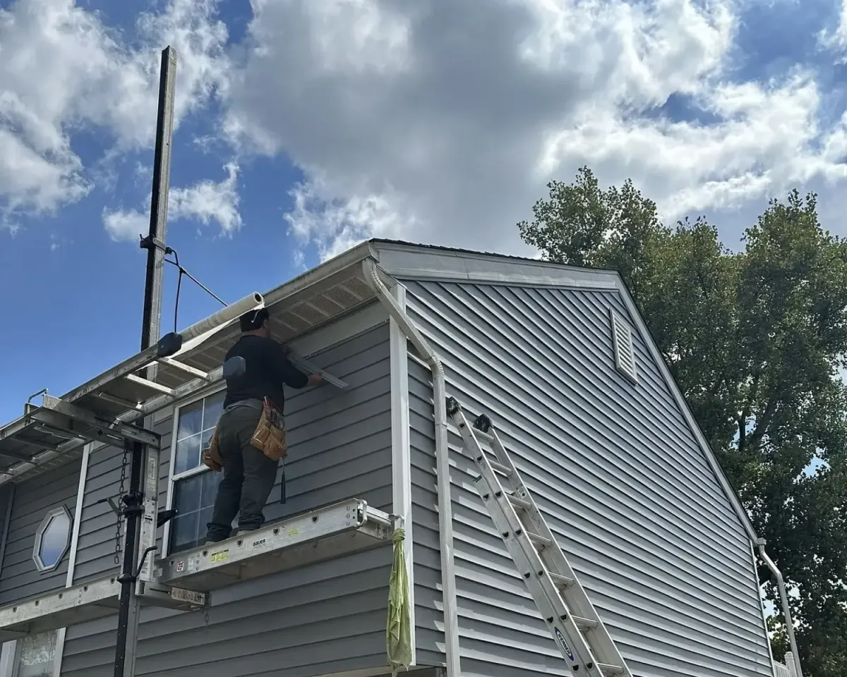 Siding contractor installing vinyl siding and trim on a two-story residential home using scaffolding and ladders under a partly cloudy sky.